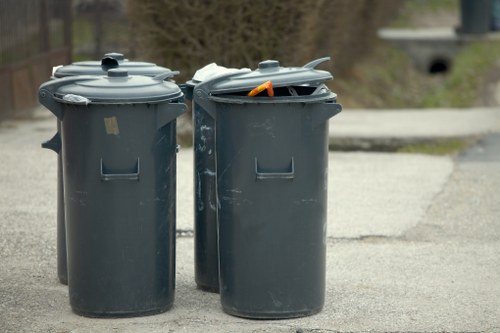 Two large grey plastic wheelie bins with closed lids are positioned on a paved surface, likely a driveway or pavement area in a residential or commercial setting. The bin on the left has a smooth, slightly shiny finish and appears to be in good condition, while the bin on the right has an open lid revealing some mixed waste inside, including an orange object that may be a piece of plastic or packaging. The background features a blurred view of what could be a garden or outdoor space, with some greenery and a concrete structure visible in the distance. The environment looks typical for waste collection, with no other visible objects or vehicles nearby. This scene relates to rubbish removal services provided by Commercial Waste Bethnal Green, reflecting typical residential or small business waste management in the Bethnal Green area. The lighting suggests daytime with overcast weather, lending a neutral and realistic tone to the image.